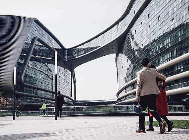 Two people walk on a boardwalk near modern glass buildings with a curved skybridge connecting them. Other pedestrians are visible in the background.