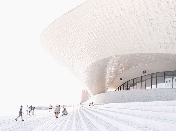 People walk near a modern, curved building with textured exterior walls and wide steps in bright daylight.