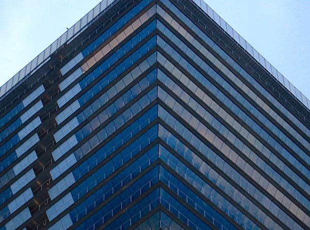 Close-up view of the corner of a modern glass office building with blue-tinted windows, showing sharp angles and reflective surfaces against a clear sky.