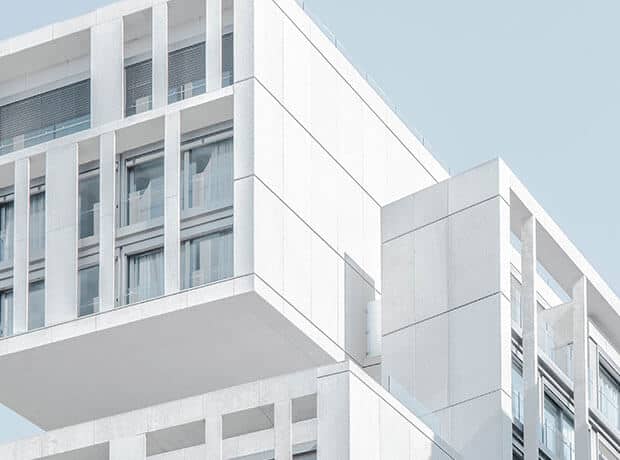 Modern white building with large glass windows and geometric architectural features against a pale blue sky.