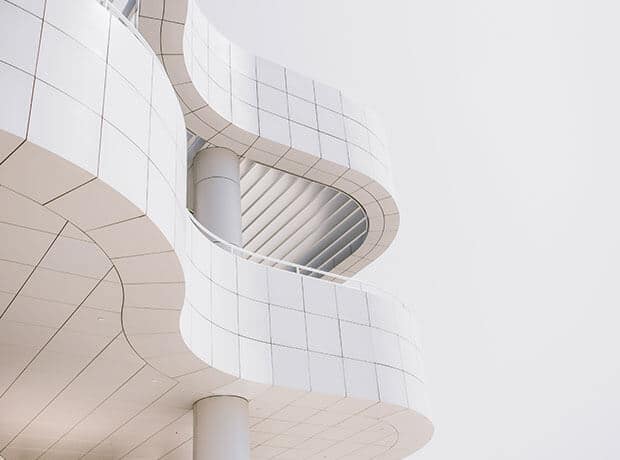Abstract view of a modern white building with curved, tiled exterior and cylindrical support columns, set against a pale sky.