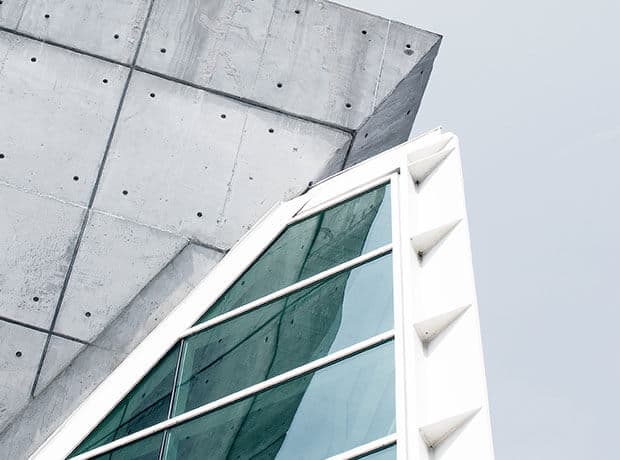 Angular modern building facade with exposed concrete, large glass windows, and white structural elements against a pale sky.