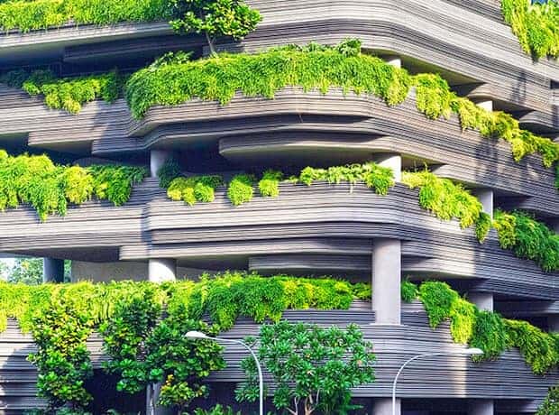 Modern multi-story building with layered horizontal lines, covered in lush green plants and trees on each level, integrating greenery into the architecture.