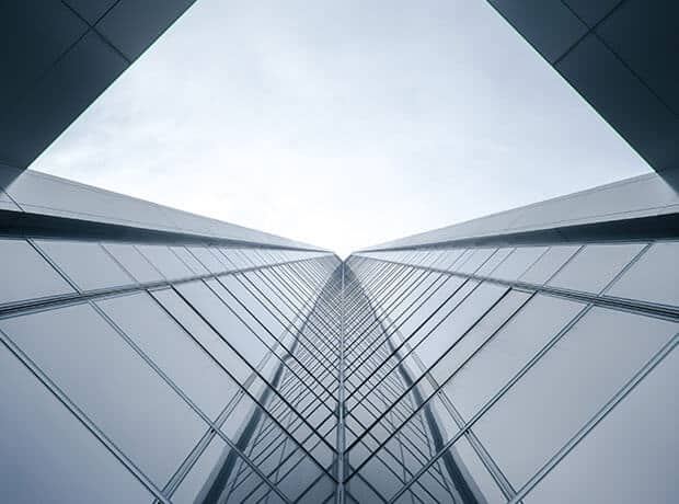 Upward view of a modern glass skyscraper with reflective windows, converging lines, and a bright sky above.